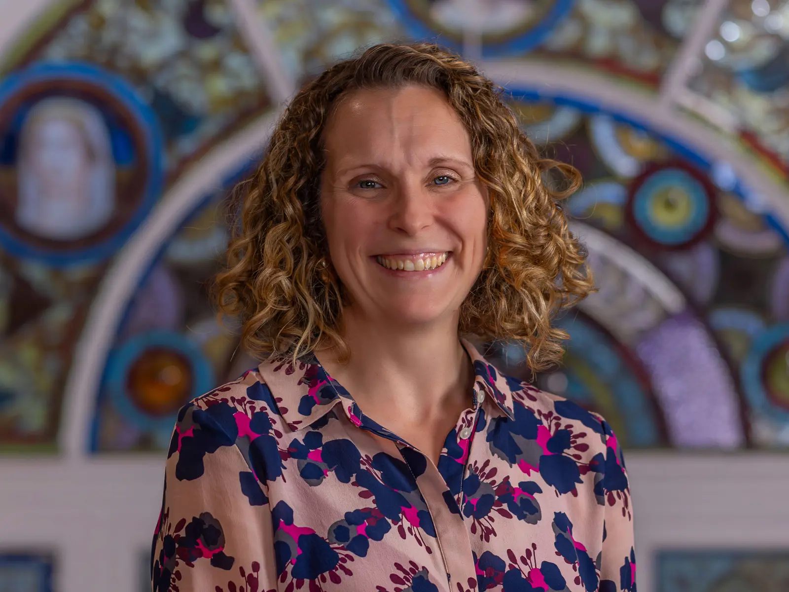 Smiling curly haired woman in a pink and purle floral blouse in front of stain glass window