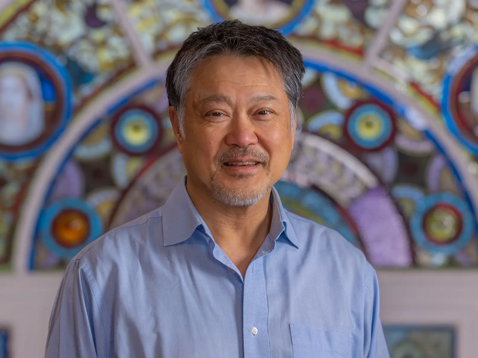 Dark haired man wearing blue shirt in front of stain glass window