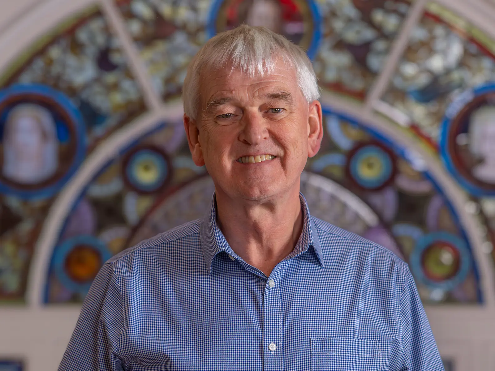 Smiling grey haired man in front of stain glass window in blue checked shirt