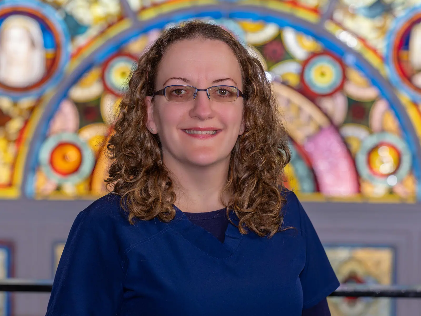 Curly haired woman in glasses and blue doctor scrubs in front of stain glass window