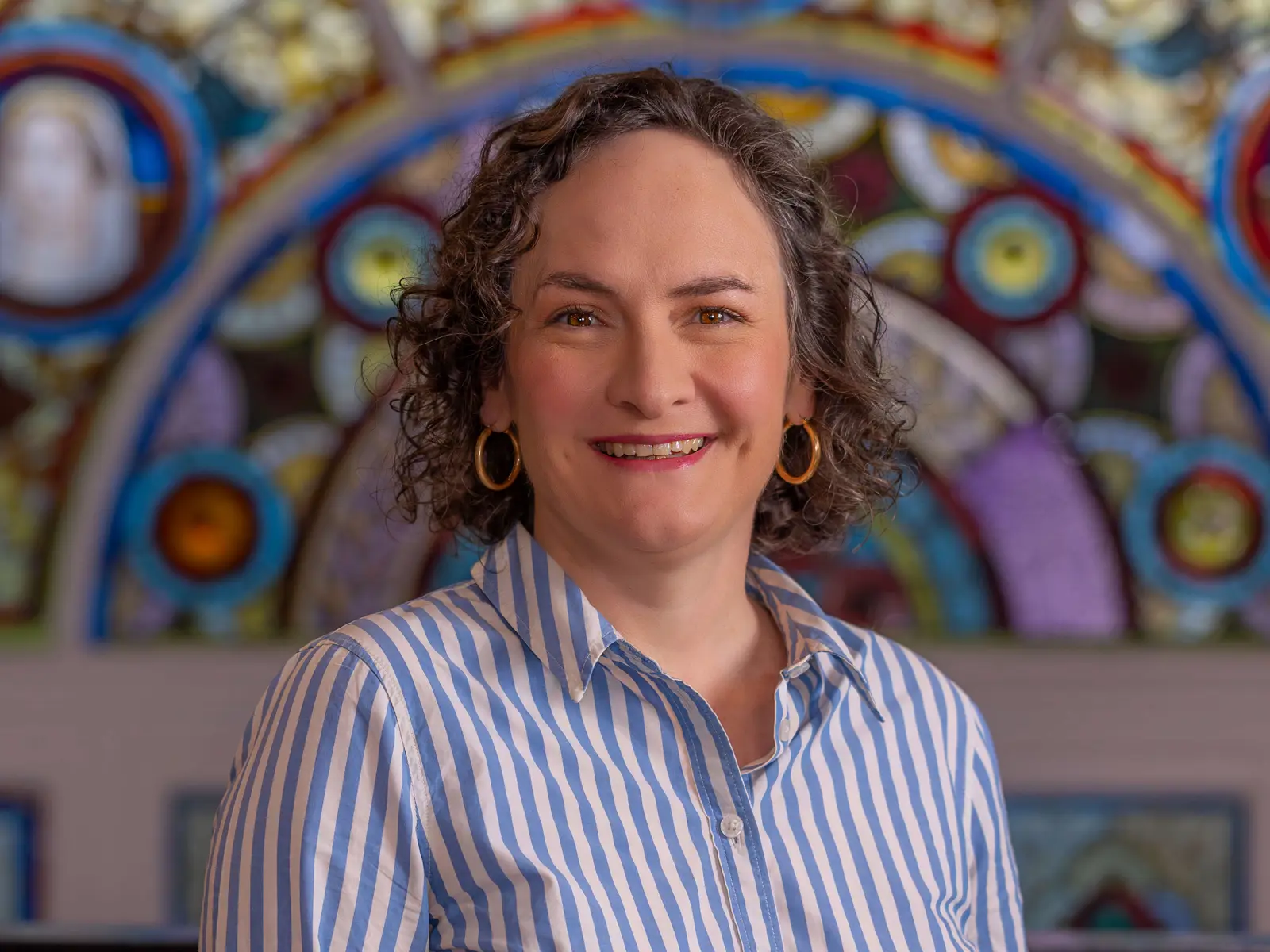 Smiling curly haired woman in blue and white shirt standing in front of stain glass window
