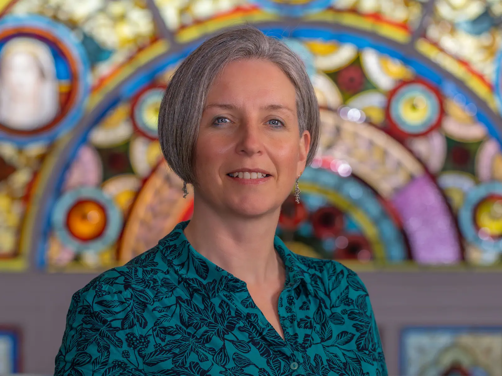 Grey short haired woman in green and black blouse in front of stain glass window