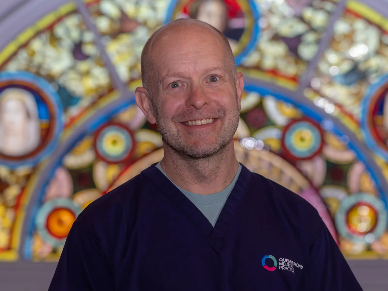 Bald smiling man in dark blue doctor scrubs in front of stain glass window